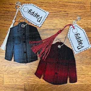 Puede incluir: Dos adornos de tela con forma de camisa abotonada, uno en degradado azul y negro y otro en rojo y negro. Cada uno cuenta con una anilla met&aacute;lica, una borla decorativa y una etiqueta de tela blanca bordada con la palabra Pappy.