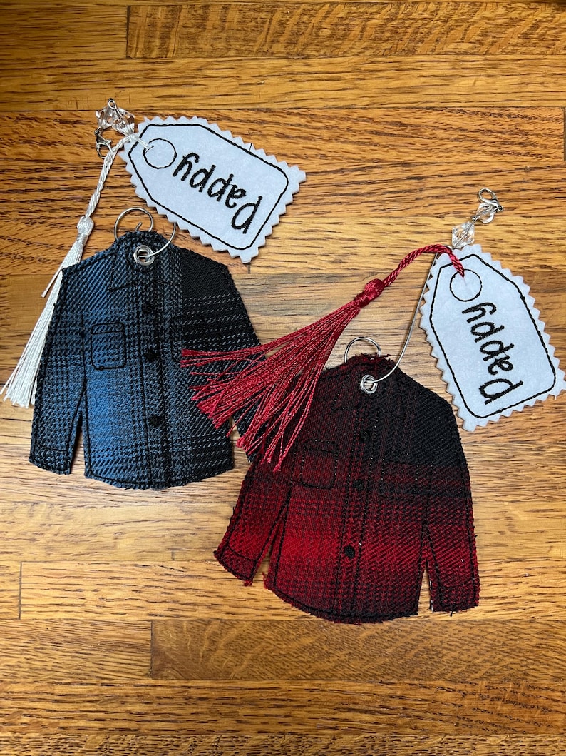 Puede incluir: Dos adornos de tela con forma de camisa abotonada, uno en degradado azul y negro y otro en rojo y negro. Cada uno cuenta con una anilla met&aacute;lica, una borla decorativa y una etiqueta de tela blanca bordada con la palabra Pappy.