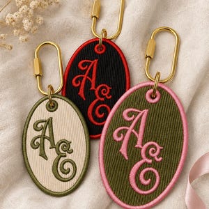 May include: Three oval-shaped embroidered keychains with gold-toned metal carabiner clips. One keychain is cream with dark green embroidery, one is black with red embroidery, and one is olive green with pink embroidery, all featuring the letters AE.
