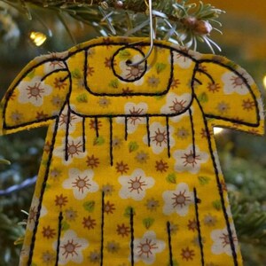Puede incluir: Un adorno de tela amarillo con forma de vestido y estampado floral blanco y marr&oacute;n. El adorno presenta contornos y l&iacute;neas verticales cosidas en negro, con un gancho de metal plateado en forma de estrella fijado en la parte superior.
