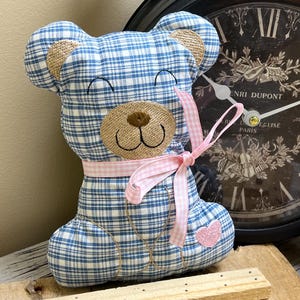 Puede incluir: Un oso de peluche hecho de tela a cuadros azul y blanca con tela texturizada color canela en las orejas y el hocico. Presenta una cinta de cuadros rosa atada al cuello y un pequeño parche de corazón rosa en la parte inferior del cuerpo.