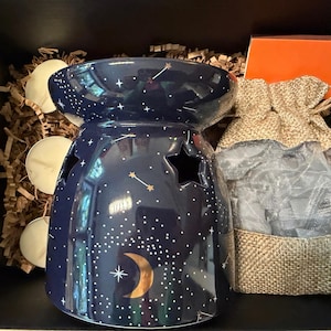 May include: A dark blue ceramic wax warmer featuring a gold crescent moon, white star patterns, and star-shaped cutouts. The set includes three white tea light candles and a burlap drawstring bag with a clear window, all nestled in shredded paper.