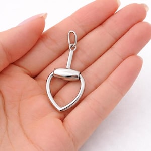 May include: A silver-colored pendant in the shape of a horse bit. The pendant has a rounded center piece and a heart-shaped bottom. The pendant is held in a person's open hand.
