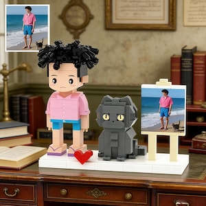 Puede incluir: Una figura de ladrillo de una persona con una camisa rosa y pantalones cortos azules, con un gato negro y una pintura de la persona en una playa. Un corazón rojo se coloca frente a la figura. La escena está sobre una base blanca.