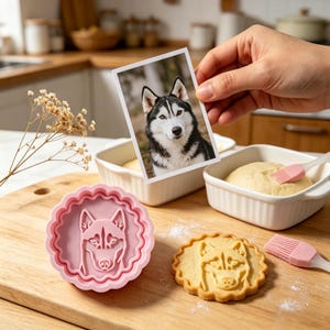 Puede incluir: Un cortador de galletas redondo rosa con un diseño de perro husky, junto a una galleta horneada con el mismo diseño. Una foto de un perro husky se sostiene sobre el cortador. Utensilios de horneado en el fondo.