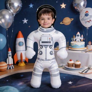 Op de afbeelding: Een opblaasbaar wit astronautenkostuum met grijze accenten en een zwarte helmuitsnede. Het pak is voorzien van een bedrukt bedieningspaneel op de borst met knoppen en wijzerplaten. Het ontwerp is bedoeld om te dragen, waarbij het gezicht door de helmopening verschijnt, wat de indruk wekt van een ruimtevaarder in een volledig drukpak.