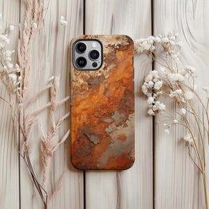 May include: A phone case with a weathered, rusty metal design. The case features shades of orange, brown, and gray, mimicking the look of aged iron. The case has a camera cutout and is surrounded by dried flowers on a light wood surface.