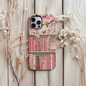 May include: A phone case featuring a bookshelf design with pink books. The top of the case has a floral pattern with pink blossoms and a butterfly. The case is decorated with a light pink and beige color scheme.