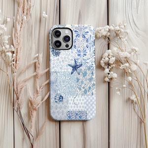 May include: A phone case with a blue and white patchwork design. The case features floral, seashell, and starfish patterns. The case is set against a wooden background, with dried flowers on either side.