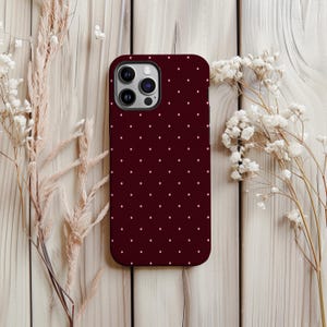 May include: A burgundy phone case featuring a pattern of small, white polka dots. The case has a camera cutout. The image shows the case on a light wood surface, with dried flowers.