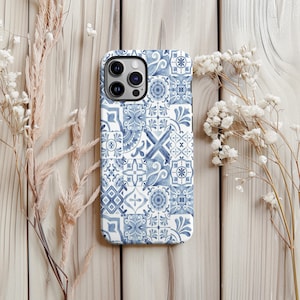 Puede incluir: Una funda de teléfono con estampado azul y blanco con un diseño de patchwork de varios patrones de azulejos. La funda tiene un acabado brillante. La funda se muestra sobre una superficie de madera clara con flores secas.