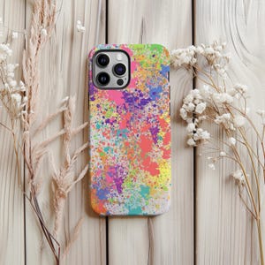 Puede incluir: Una funda de teléfono colorida con un diseño vibrante de pintura salpicada. La funda presenta una mezcla de colores brillantes, incluyendo rosa, amarillo, verde y morado. La funda se muestra sobre una superficie de madera clara.