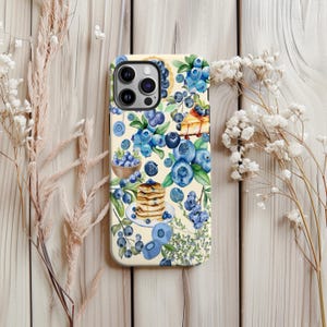 Puede incluir: Funda de teléfono con fondo crema con un diseño de acuarela de arándanos, panqueques y tarta. El diseño incluye bayas azules, hojas verdes y un cuenco de madera. La funda tiene un acabado brillante.
