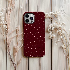 May include: A maroon phone case featuring a pattern of small white hearts. The case has a camera cutout. It is displayed with dried flowers on a wooden surface. The case is designed for a smartphone.