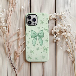 May include: A light green phone case featuring a large bow design in the center, surrounded by smaller bow illustrations. The case has a matte finish and is decorated with small heart shapes.