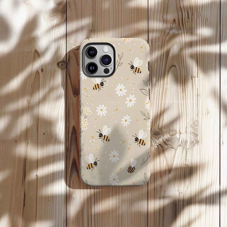 May include: A beige phone case decorated with bees and daisies. The bees have black and yellow stripes and white wings. The daisies are white with yellow centres. The case is on a wooden surface.