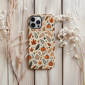 Può includere: Una custodia per telefono con un design a tema autunnale con zucche, foglie e fogliame nei toni dell'arancione, marrone e verde su uno sfondo crema. La custodia è circondata da fiori secchi su una superficie di legno chiaro.