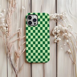 May include: A green and light green checkered phone case. The case is rectangular with rounded corners and a camera cutout. The background is a light wooden surface with dried flowers.