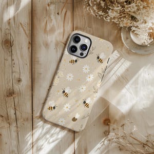 May include: Beige phone case with a pattern of cartoon bees, white daisies, and yellow dots. The case has a camera cutout and is on a wooden surface. The design has a whimsical, nature-inspired theme, with a light, airy aesthetic.