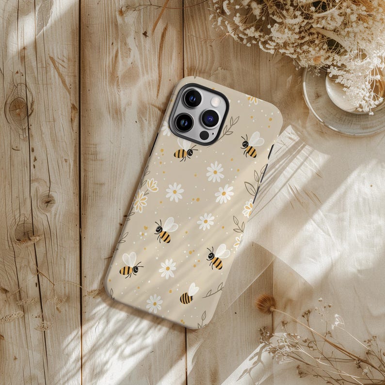 May include: Beige phone case with a pattern of cartoon bees, white daisies, and yellow dots. The case has a camera cutout and is on a wooden surface. The design has a whimsical, nature-inspired theme, with a light, airy aesthetic.