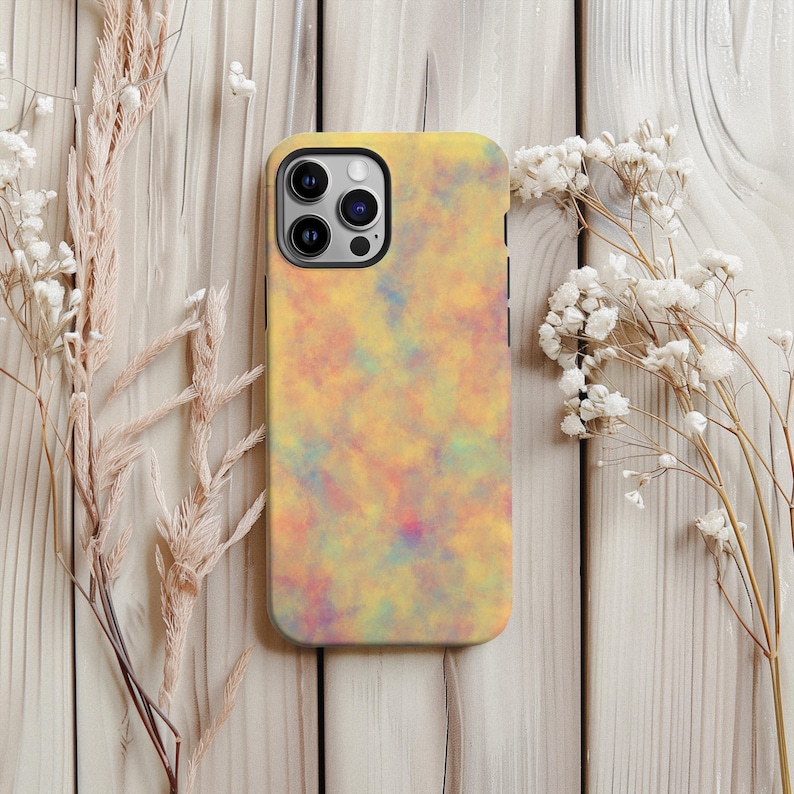 May include: A yellow phone case with a colorful, abstract design. The case features a pattern of blended colors, including yellow, orange, pink, and blue. The phone case is shown on a light wooden surface, with dried flowers.