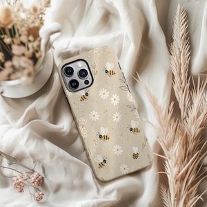 May include: A beige phone case decorated with bees, white daisies, and yellow dots. The case has a glossy finish and a camera aperture. The design includes small green leaves and a floral border.