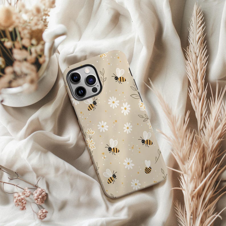 May include: A beige phone case decorated with bees, white daisies, and yellow dots. The case has a glossy finish and a camera aperture. The design includes small green leaves and a floral border.