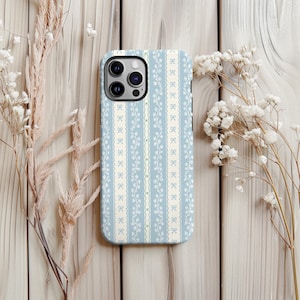 Pode incluir: Uma capa de telefone azul claro com um padrão repetido de laços brancos e vinhas florais. A capa tem um design de listras verticais com uma borda creme semelhante a renda. A capa do telefone está em uma superfície de madeira clara com flores secas.