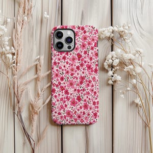 Puede incluir: Una funda de tel&eacute;fono rosa floral con un patr&oacute;n de peque&ntilde;as flores rosas sobre un fondo blanco. La funda tiene un borde negro y un recorte para la c&aacute;mara. La funda est&aacute; sobre una superficie de madera clara, con flores secas a ambos lados.