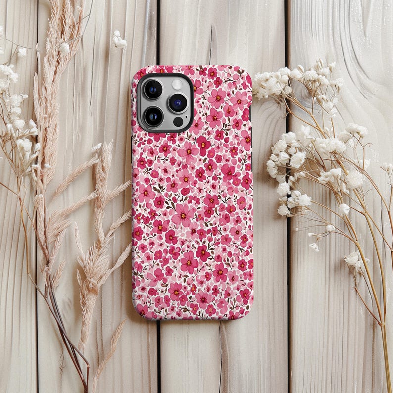 Puede incluir: Una funda de tel&eacute;fono rosa floral con un patr&oacute;n de peque&ntilde;as flores rosas sobre un fondo blanco. La funda tiene un borde negro y un recorte para la c&aacute;mara. La funda est&aacute; sobre una superficie de madera clara, con flores secas a ambos lados.