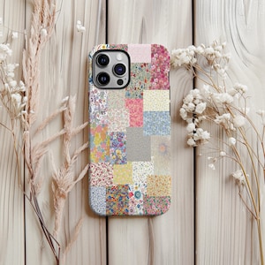 May include: A floral patchwork phone case featuring a variety of colorful floral patterns in a grid-like design. The case has a black border and a camera cutout. The background is a light wooden surface with dried flowers.