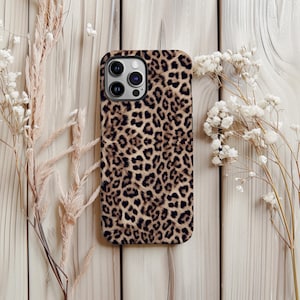 May include: A phone case with a leopard print design. The case features a pattern of brown and black spots on a tan background. The phone case is surrounded by dried flowers and placed on a light wood surface.