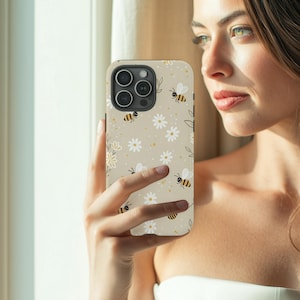 May include: A beige phone case with a pattern of white daisies, yellow bees, and green leaves. The case has a black camera module. The phone case is held by a person.