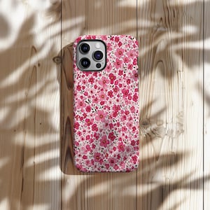 Puede incluir: Una funda de tel&eacute;fono floral con un patr&oacute;n de peque&ntilde;as flores rosas sobre un fondo blanco. La funda tiene un m&oacute;dulo de c&aacute;mara negro y est&aacute; colocada sobre una superficie de madera con sombras de hojas.