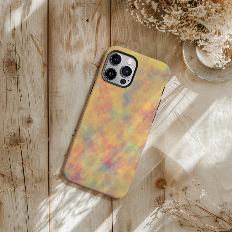 May include: A smartphone case with a colorful, abstract design. The case features a pattern of blended yellow, orange, pink, and blue hues. The phone case has a black border and a camera cutout. The case is lying on a wooden surface.