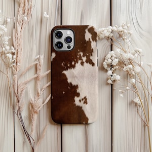 Peut inclure: Coque de téléphone marron et blanche à motif peau de vache. L'étui a une finition brillante et une découpe pour l'appareil photo. Entouré de fleurs séchées sur un fond en bois clair.