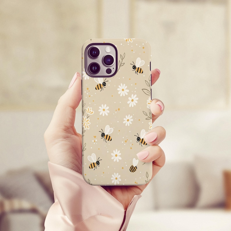 May include: A beige phone case with a repeating pattern of cartoon bees and white daisies. The case has a matte finish and a protective design. The phone case is designed to fit a smartphone.