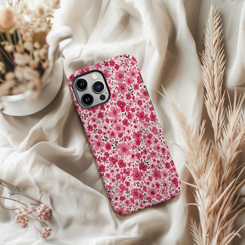 Puede incluir: Una funda de tel&eacute;fono con un dise&ntilde;o floral. La funda presenta un dise&ntilde;o repetido de peque&ntilde;as flores rosas sobre un fondo blanco. La funda del tel&eacute;fono se muestra sobre una tela blanca.