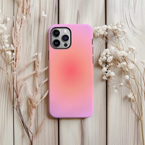 Puede incluir: Una funda de teléfono rosa con un diseño degradado, que transiciona de un centro coral vibrante a un borde exterior rosa suave. La funda se muestra sobre una superficie de madera clara, con flores secas a ambos lados.
