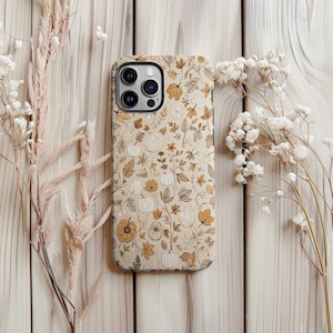 May include: A beige phone case with a floral pattern featuring pumpkins, leaves, and flowers in shades of brown, orange, and white. The case is surrounded by dried flowers and set against a light wood background.