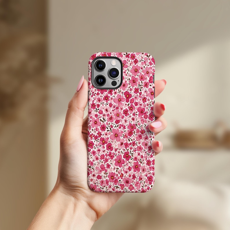 Puede incluir: Una funda de tel&eacute;fono floral con un patr&oacute;n de peque&ntilde;as flores rosas sobre un fondo blanco. La funda se sostiene en una mano con esmalte de u&ntilde;as rojo. La funda del tel&eacute;fono tiene un m&oacute;dulo de c&aacute;mara con tres lentes.