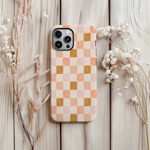 May include: A phone case with a checkerboard pattern in shades of peach, pink, and brown. The case has a smooth finish and a camera cutout. The design is a modern, abstract style. The case is surrounded by dried flowers.
