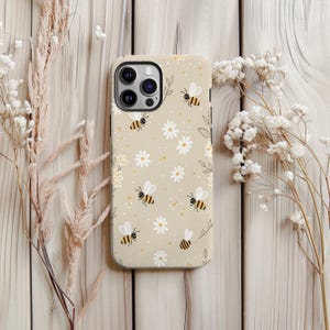 May include: A beige phone case with a pattern of bees, daisies, and small yellow dots. The case has a matte finish and a camera cutout. The background is a light wooden surface with dried flowers.