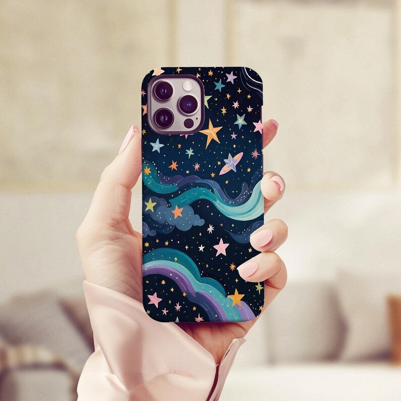 May include: A phone case with a whimsical night sky design. The case features a dark blue background with colorful stars, clouds, and a rainbow. The design includes yellow, pink, and blue hues, creating a dreamy aesthetic. The case is held in a hand.