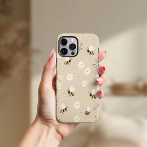 May include: A beige phone case with a repeating pattern of cartoon bees and white daisies. The case has a matte finish and is designed for a smartphone. The design includes small yellow dots and green leaf accents.