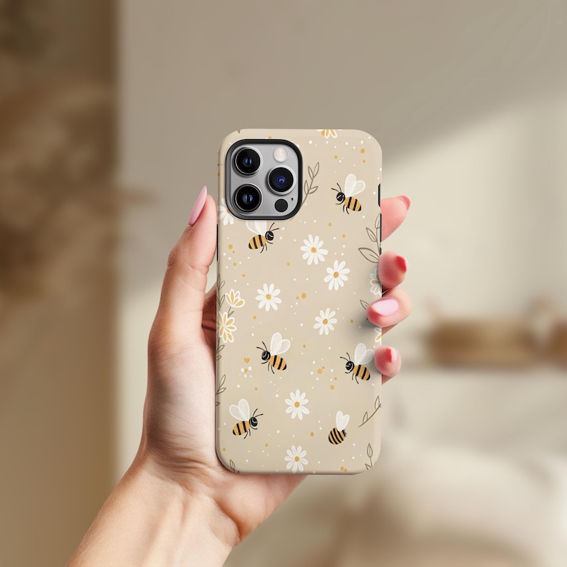 May include: A beige phone case with a repeating pattern of cartoon bees and white daisies. The case has a matte finish and is designed for a smartphone. The design includes small yellow dots and green leaf accents.