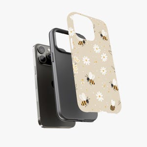 May include: A phone case with a beige background featuring a pattern of bees and white daisies. The case is shown in three parts: the phone, a black bumper, and the patterned cover. The bees have black and yellow stripes.