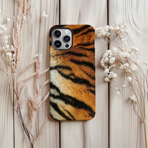 May include: A smartphone case featuring a tiger stripe pattern in shades of orange, brown, and black. The case has a glossy finish and a camera cutout. The background is a light-colored wooden surface with dried flowers.