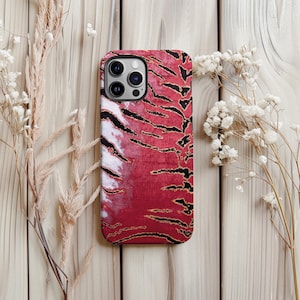 May include: A phone case featuring a red and black tiger stripe design. The case has a glossy finish and a camera cutout. The design includes black stripes over a red background, with gold accents. The case is displayed on a light wood surface.