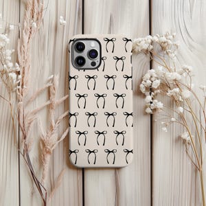 May include: A beige iPhone case with a repeating pattern of black bow tie illustrations. The case has a glossy finish. The phone case is surrounded by dried flowers on a light wooden surface.